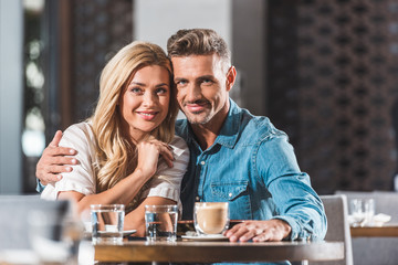 happy tender heterosexual couple hugging at table in cafe and looking at camera