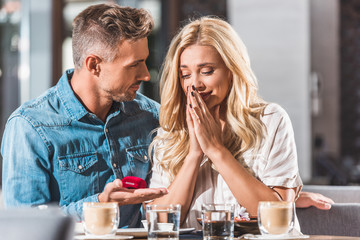 handsome boyfriend making proposal to surprised girlfriend and holding ring box in cafe