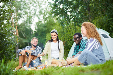 Fototapeta premium Young happy campers relaxing by campfire, talking and frying marshmellows on backpack trip