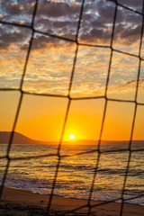 Fototapeta premium View of the sunrise through the volleyball net. Early morning , dramatic sunrise over sea and mountain.