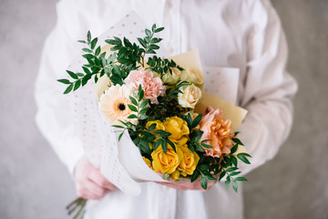 Very nice young man in a white shirt holding blossoming flower bouquet of  fresh gerbera, roses, carnations in yellow and peach colors on the grey wall background