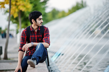 Young indian student man at checkered shirt and jeans with backpack posed on evening city against fountains.