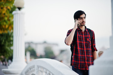 Young indian student man at checkered shirt and jeans with backpack speaking on mobile phone.