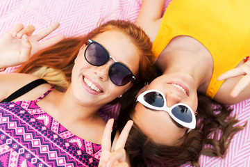 summer fashion, eyewear and people concept - smiling teenage girls in sunglasses lying on picnic blanket and showing peace hand sign