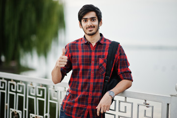 Young indian student man at checkered shirt and jeans with backpack posed on evening city against...