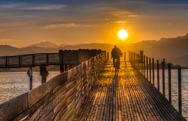 Obraz premium Biking during Dramatic sunrise during the Fall Equinox over the historical holzsteg pedestrian bridge crossinhg the Upper Zurich Lake (Obersee), part of the old Way of Saint James, Switzerland