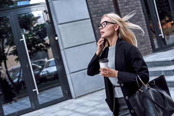 Young stylish woman talking by smartphone. She walks with morning coffee