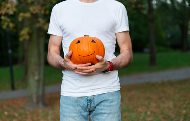 Handsome brunette man in white t-shirt holding orange pumpkin with drawing happy face and posing at...