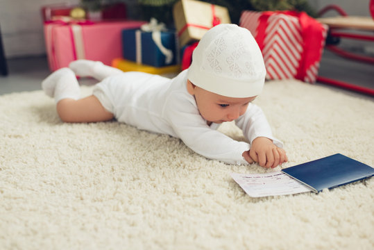 cute little baby with flight ticket and passport lying on floor with christmas gifts blurred on background