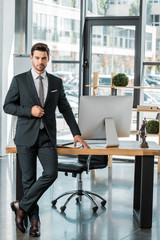 handsome businessman standing near table in office and looking at camera