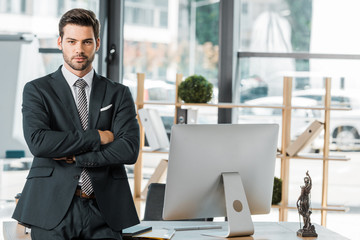 Fototapeta premium handsome businessman standing with crossed arms near table in office