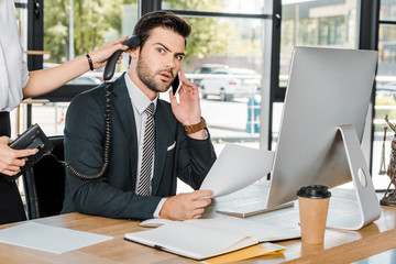 shocked businessman talking by smartphone and secretary holding handset of stationary telephone near his ear