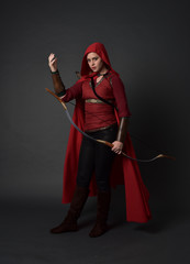 full length portrait of brunette girl wearing red medieval costume and cloak, holding a bow and...