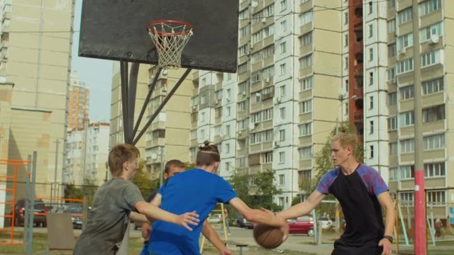 Rear view of teenage basketball player bouncing ball and making field goal attempt in the paint with resistance of defenders while playing streetball game on outdoor court over cityscape background.