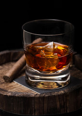 Glass of whiskey with ice cubes and cigar on top of wooden barrel. Cognac brandy drink