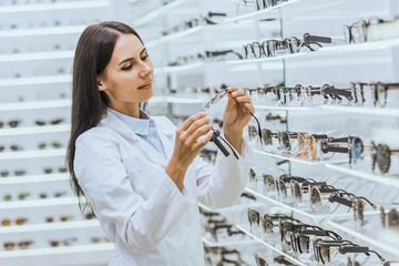 beautiful optometrist looking at eyeglasses near shelves in ophthalmic shop