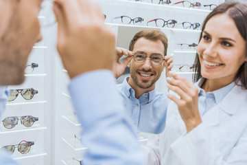 cheerful female optometrist holding mirror while smiling man choosing eyeglasses in ophthalmic shop - obrazy, fototapety, plakaty