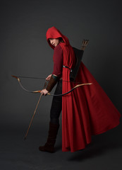 full length portrait of brunette girl wearing red medieval costume and cloak, holding a bow and...