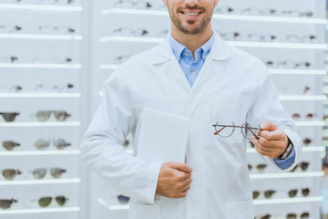 cropped view of optometrist in white coat holding eyeglasses and digital tablet in optica