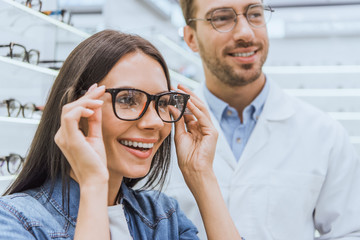 portrait of smiling woman choosing eyeglasses while male optometrist standing near in optics