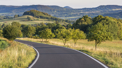 The road leading down. A view of the countryside before the road. Landscape lit by the setting sun.