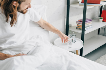 selective focus of young bearded man with long hair having headache and taking glass of water in bedroom at home