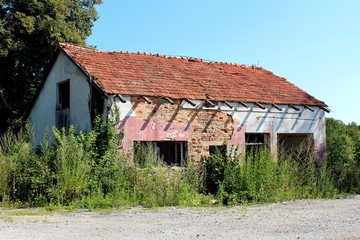 Fototapeta na wymiar Abandoned small store red brick building destroyed in war with missing windows and doors, broken roof without some tiles, visible wooden beams without gutter completely surrounded with trees, high unc
