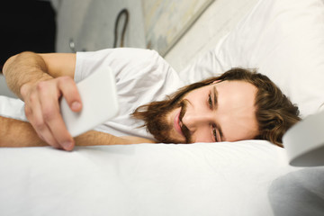 Obraz na płótnie Canvas smiling bearded man with long hair using smartphone in bed during morning time at home