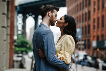 Love story in New York. Gorgeous couple of American man with beard and tender Eastern woman hug each other before the cityscape of Brooklyn bridge somewhere in New York