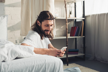 happy Jesus in crown of thorns using smartphone in bedroom at home