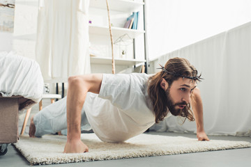selective focus of Jesus in crown of thorns doing push ups during morning time in bedroom at home