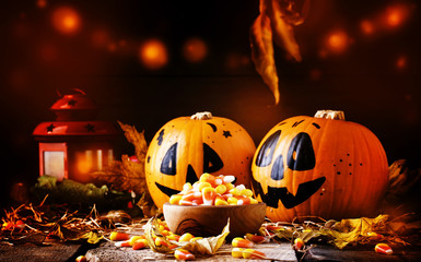 Halloween festive composition with sweet corn in bowl and smiling pumpkins guards, lantern, straw and fallen leaves on dark wooden background, rustic style, selective focus