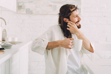side view of happy Jesus in earphones listening music and holding cup of coffee in kitchen at home