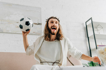 happy Jesus in crown of thorns sitting on sofa with soccer ball and watching football match at home
