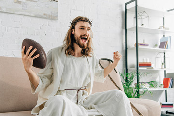 excited Jesus in crown of thorns gesturing by hand and watching american football on sofa at home