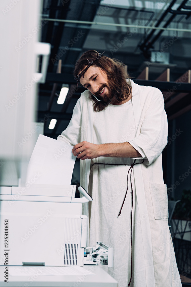 Wall mural cheerful jesus in crown of thorns and robe talking on smartphone and using copy machine in modern of