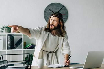 aggressive Jesus in crown of thorns and robe writing in textbook and pointing by finger at table with laptop in modern office - obrazy, fototapety, plakaty