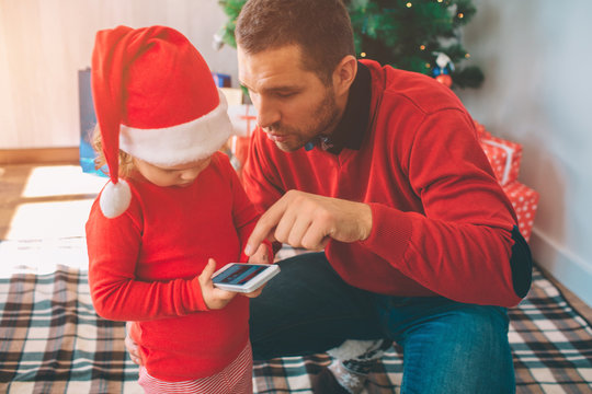 Merry Christmas and Happy New Year. Picture of father and daughter together. Kid holds phone in hands. Man shows and points on technology. Child wears Christmas hat. They are at tree with boxes.