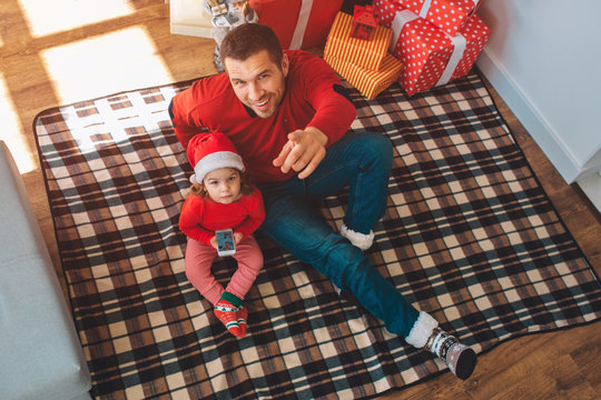 Merry Christmas and Happy New Year. Young dad sits on blanket with child and look on camera. He smiles and points on that. Kid look up as well. She holds phone in hands. They are together.