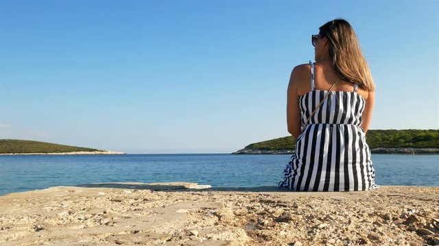 Young caucasian woman sitting alone on stone dock on Croatian coast and looking at blue sea