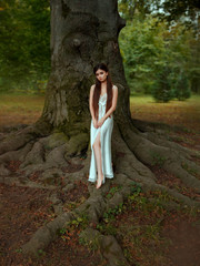 Young princess with long hair in a white, vintage, nightdress. The girl stands on the huge roots of an ancient tree. Fantastic art photo.