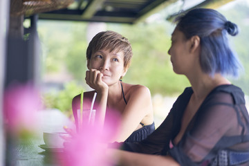 portrait of 2 asian women chatting, drinking & smiling at beach bar in summer