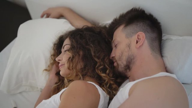 a young girl wakes up in the arms of a beloved bearded man. the happy curly-haired woman smiles. The little dog Jack Russell Terrier is lying side by side on the bed in a fluffy white blanket. 4k