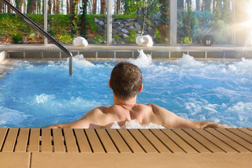Handsome man in hot tub spa in luxurious hotel with big glass windows with nature view.