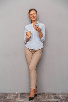 Young woman posing isolated over grey wall background using mobile phone drinking coffee.