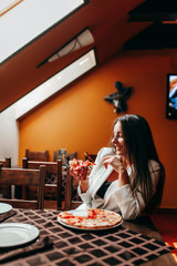 Beautiful stylish brunette girl in white jacket eating pizza at restaurant.