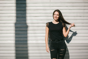 A brunette female with long hair wearing a blank black t-shirt i while standing on a concrete wall background on a street. Empty space for text or design.