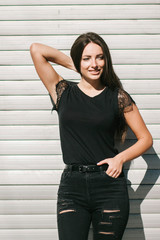 A brunette female with long hair wearing a blank black t-shirt i while standing on a concrete wall background on a street. Empty space for text or design.