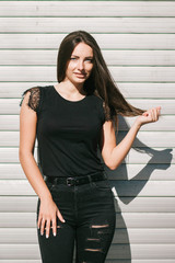 A brunette female with long hair wearing a blank black t-shirt i while standing on a concrete wall background on a street. Empty space for text or design.