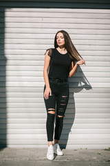 A brunette female with long hair wearing a blank black t-shirt i while standing on a concrete wall background on a street. Empty space for text or design.